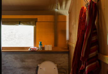 Binnenzicht van een safaritentbadkamer met wit toilet, handdoeken, vuilbak en een raam.