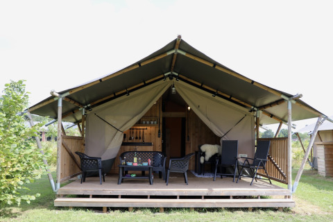 Tenda safari con servizi igienici, veranda in legno e sedie all'aperto, immersa nel verde.