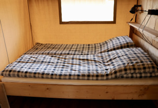 Vista interna di un letto con biancheria a quadri all’interno di una tenda safari al Campeggio Heerlijkheid Vorenseinde.