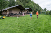 Children running on grass near a safari tent with toilet, surrounded by toys and lush green trees.