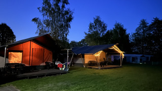Aftenbillede af en oplyst safaritelt med toilet på Campingplads Emmen, Holland, omgivet af træer.