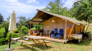 Outdoor view of a safari tent with wooden seating area, hammock and trees under a partly cloudy sky.