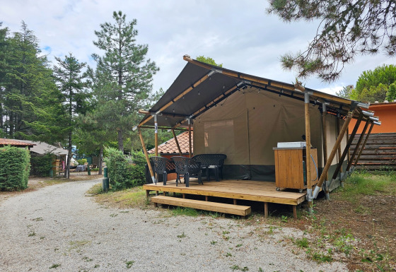 Safari tent on a campsite, featuring a wooden deck with plastic chairs, surrounded by green trees.