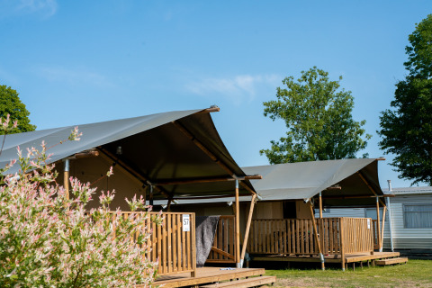Safari-Zelte mit Terrasse und sanitären Anlagen, umgeben von Bäumen und blauem Himmel.