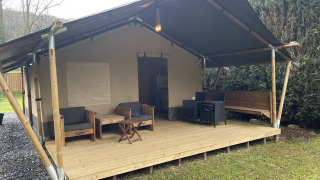 Tenda safari con veranda in legno coperta e area salotto con mobili all'aperto immersa nel verde.