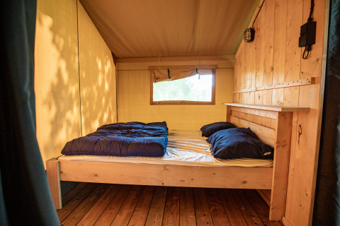 Gezellige slaapkamer in een safaritent met houten bed, blauwe dekens, raam en canvas zijwanden.