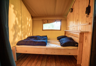 Gezellige slaapkamer in een safaritent met houten bed, blauwe dekens, raam en canvas zijwanden.