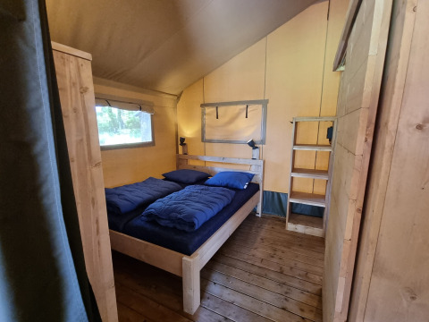 Interieur van een safaritent met een tweepersoonsbed, blauwe dekbedden en houten meubels op houten vloer.