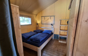 Interieur van een safaritent met een tweepersoonsbed, blauwe dekbedden en houten meubels op houten vloer.