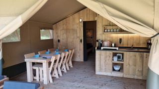 Binnenkant van een safaritent met houten tafel, stoelen, keukenblok en een sfeervolle kampeeruitstraling.