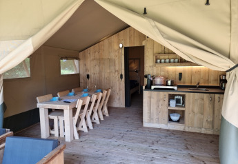 Binnenkant van een safaritent met houten tafel, stoelen, keukenblok en een sfeervolle kampeeruitstraling.