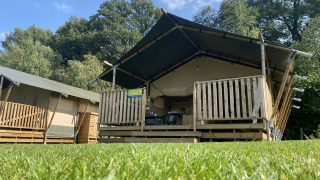 Safari tent with private porch and sanitary facilities on a grass lawn under blue sky and trees.