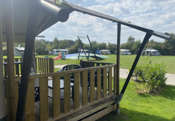 Blick von einem Safari-Zelt mit Veranda auf Campingplatz Dal van de Mosbeek, Niederlande, mit Wohnwagen und Spielplatz.