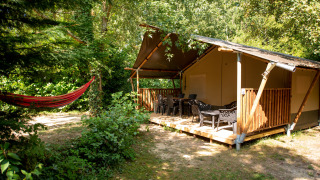 Safari tent with a wooden deck, outdoor chairs, and a red hammock set in a lush green forest.