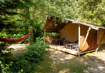 Safari tent with a wooden deck, outdoor chairs, and a red hammock set in a lush green forest.