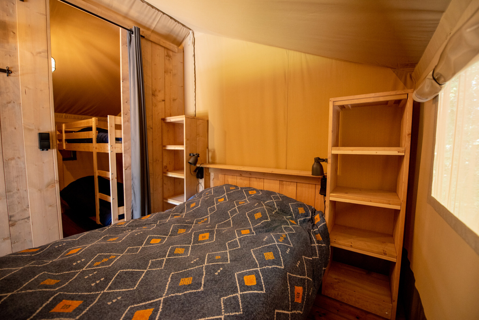 Binnenkant van een safaritent met tweepersoonsbed, stapelbedden, houten planken en zacht daglicht.