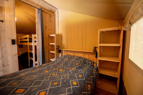 Interior of a safari tent featuring a double bed, bunk beds, wooden shelves, and soft natural light.