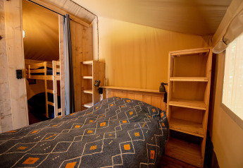 Interior of a safari tent featuring a double bed, bunk beds, wooden shelves, and soft natural light.