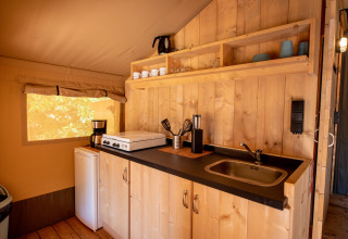 Compact kitchenette inside a safari tent, featuring a sink, cooktop, fridge, and open wooden shelves.