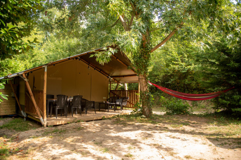 Tenda safari con bagno privato nel bosco, ampia veranda, sedie di plastica e amaca all’aperto.