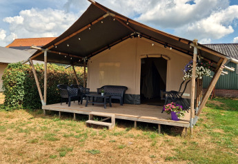 Safari tent with a wooden veranda, outdoor seating and flowers, set on a grassy area on a cloudy day.