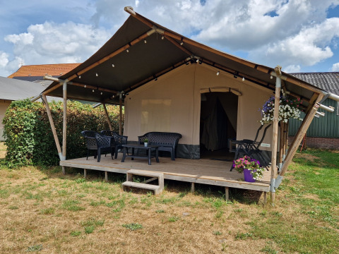 Safari telt med veranda, havemøbler og blomsterkrukker, placeret på en græsplæne under en skyet himmel.