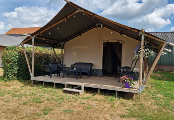 Safari telt med veranda, havemøbler og blomsterkrukker, placeret på en græsplæne under en skyet himmel.