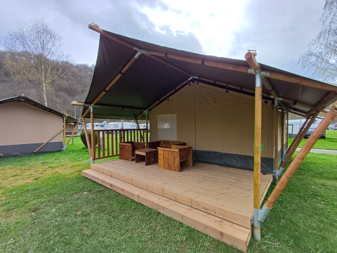 Safari-Zelt mit Holzveranda und Sitzgruppe auf einer grünen Wiese unter bewölktem Himmel.