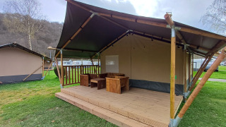 Tenda safari con veranda in legno e sedute all’aperto su prato verde sotto un cielo nuvoloso.