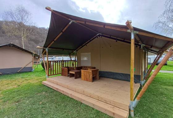 Safari-Zelt mit Holzveranda und Sitzgruppe auf einer grünen Wiese unter bewölktem Himmel.