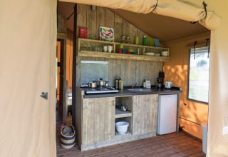 Køkkenområde i en safaritelte med træpaneler, gasblus, køleskab og hylder med service og køkkenudstyr.