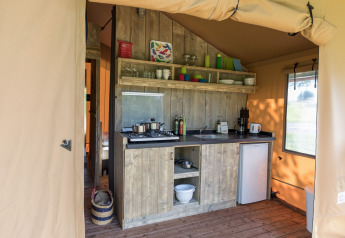 Coin cuisine d'une tente safari avec placards en bois, réchaud, évier, frigo, vaisselle et étagères.