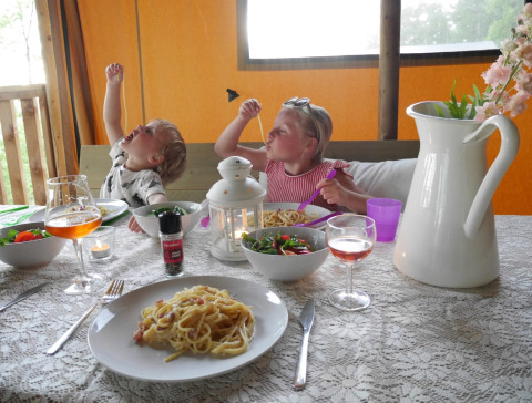 Due bambini mangiano spaghetti con allegria a un tavolo in una tenda safari, decorazioni estive intorno.