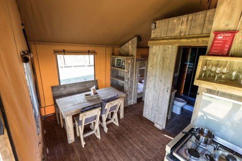 Binnenzicht van een safaritent met houten meubels, eettafel, keuken en stapelbedden op de achtergrond.