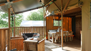 Tenda safari con veranda coperta, mobili in legno e zona pranzo, immersa tra alberi verdi nella natura.
