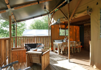 Safari telt med overdækket terrasse, træmøbler og spiseplads, omgivet af grønne træer i naturen.