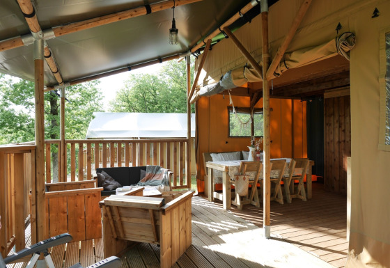 Safari tent with covered deck, wooden furniture and dining area, set among lush green trees outdoors.