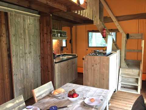 Interno di una tenda safari con arredamento in legno, cucina, zona pranzo e scala verso il soppalco.