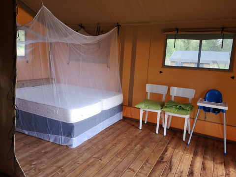 Binnenzicht van een safaritent met bed onder muskietennet, twee stoelen, kinderstoel en houten vloer.