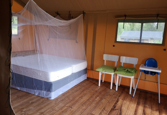 Intérieur d'une tente safari avec lit sous moustiquaire, deux chaises, chaise haute et sol en bois.