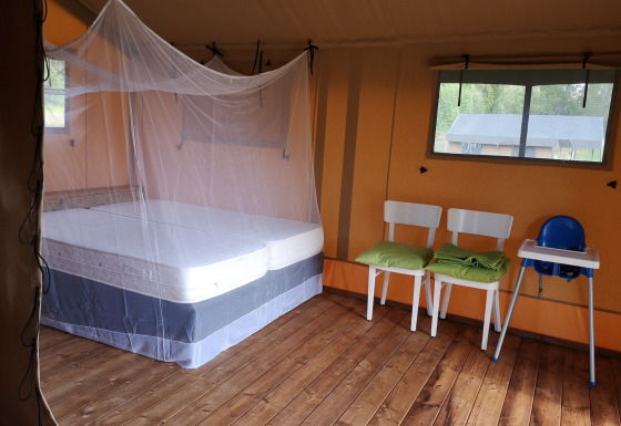 Interior of a safari tent with a bed under mosquito net, two chairs, a high chair, and wooden floor.