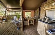 Gezellige safaritent met houten meubilair, keuken, eettafel en terras met ligzetels in het groen.