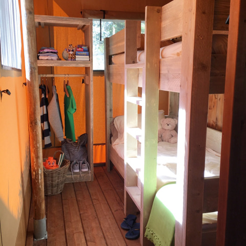 Interieur van een safaritent met houten stapelbedden, open kast met kledij en veel daglicht.