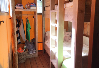 Interiør af et safaritelt med køjesenge i træ, en åben garderobe med tøj og naturlig sollys.
