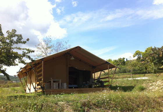 Safari tent with private sanitary facilities set on a grassy field, surrounded by trees and a blue sky.