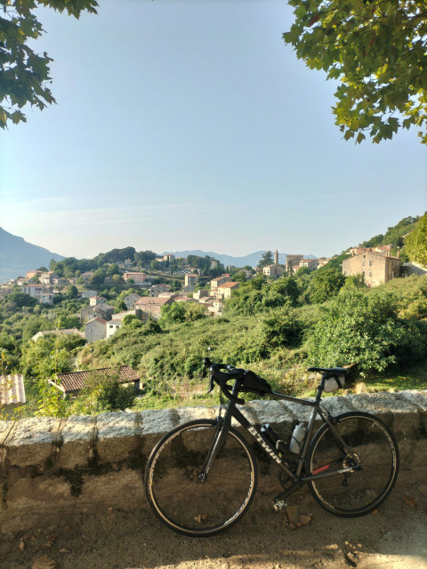 Fiets leunt tegen muur met zicht op Estang in Occitanië, Frankrijk, omringd door groene heuvels.