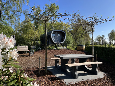 Außenansicht des Anthropod Tiny House mit Picknicktisch, Bank, Fahrrad und Bäumen im Grünen.