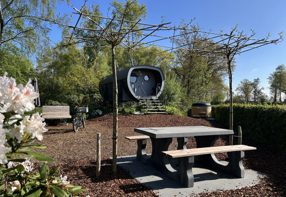 Vue extérieure de la tiny house Anthropod avec table de pique-nique, bancs, vélo et arbres autour.