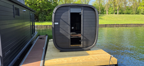 Et moderne, sort sauna-hus på flåde ved siden af en husbåd på vandet med grønne træer i baggrunden.