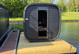 Cabina de sauna negra y moderna sobre un muelle flotante junto a una casa flotante en un lago tranquilo.